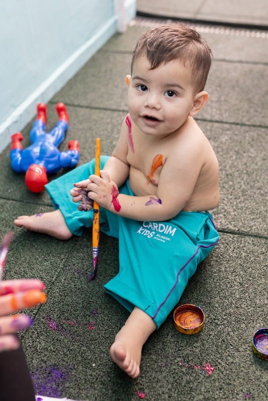 Bebê da Cardim Kids sentado no chão pintando com tinta e pincel