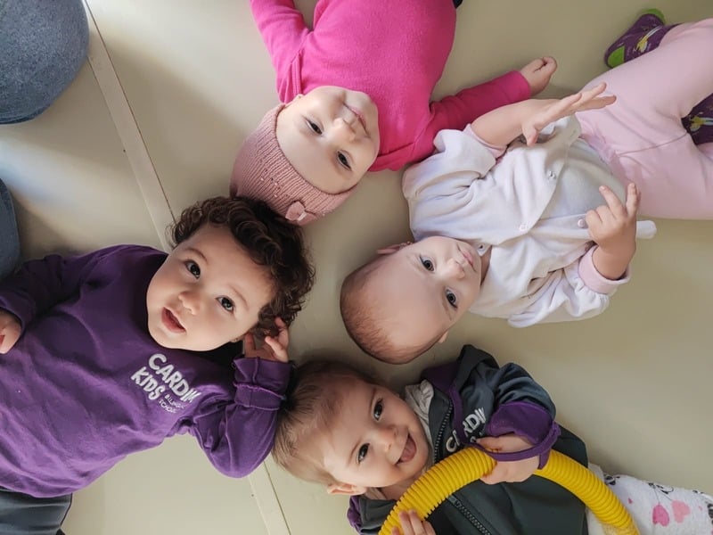 Bebês da Cardim Kids deitados em roda, de mãos dadas, com os uniformes roxos da escola